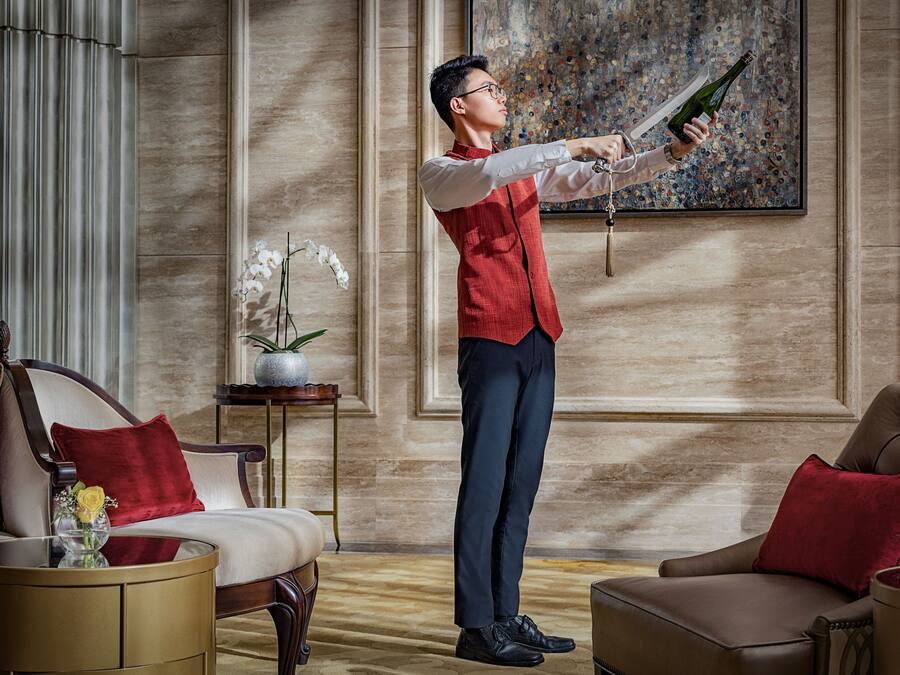 A man in a red vest and dark trousers holding a champagne bottle in a well-furnished room with a large painting on the wall, a potted plant, and a sofa with red cushions.