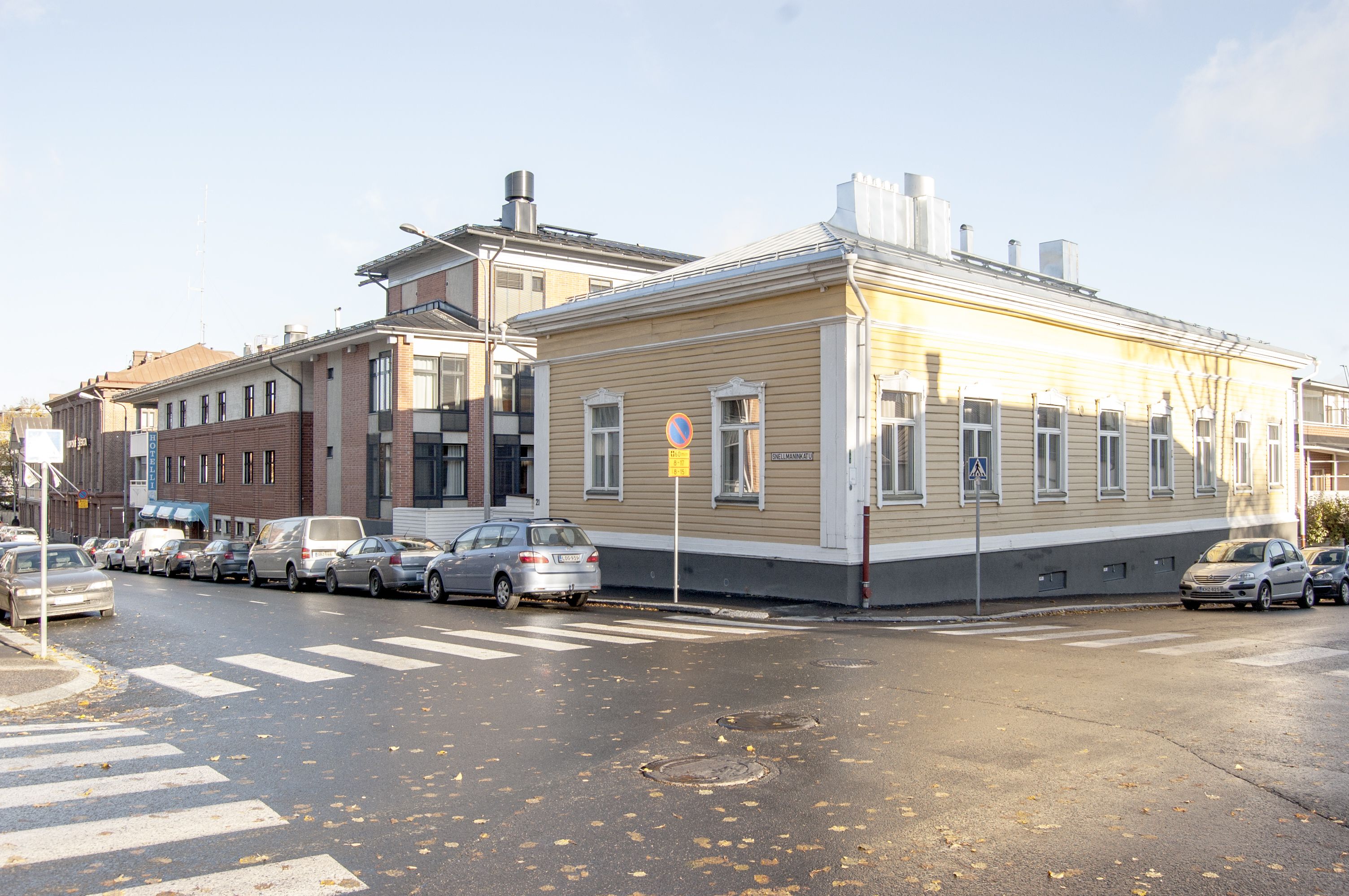 A street view with parked cars, a pedestrian crossing, and buildings with a mix of architectural styles.