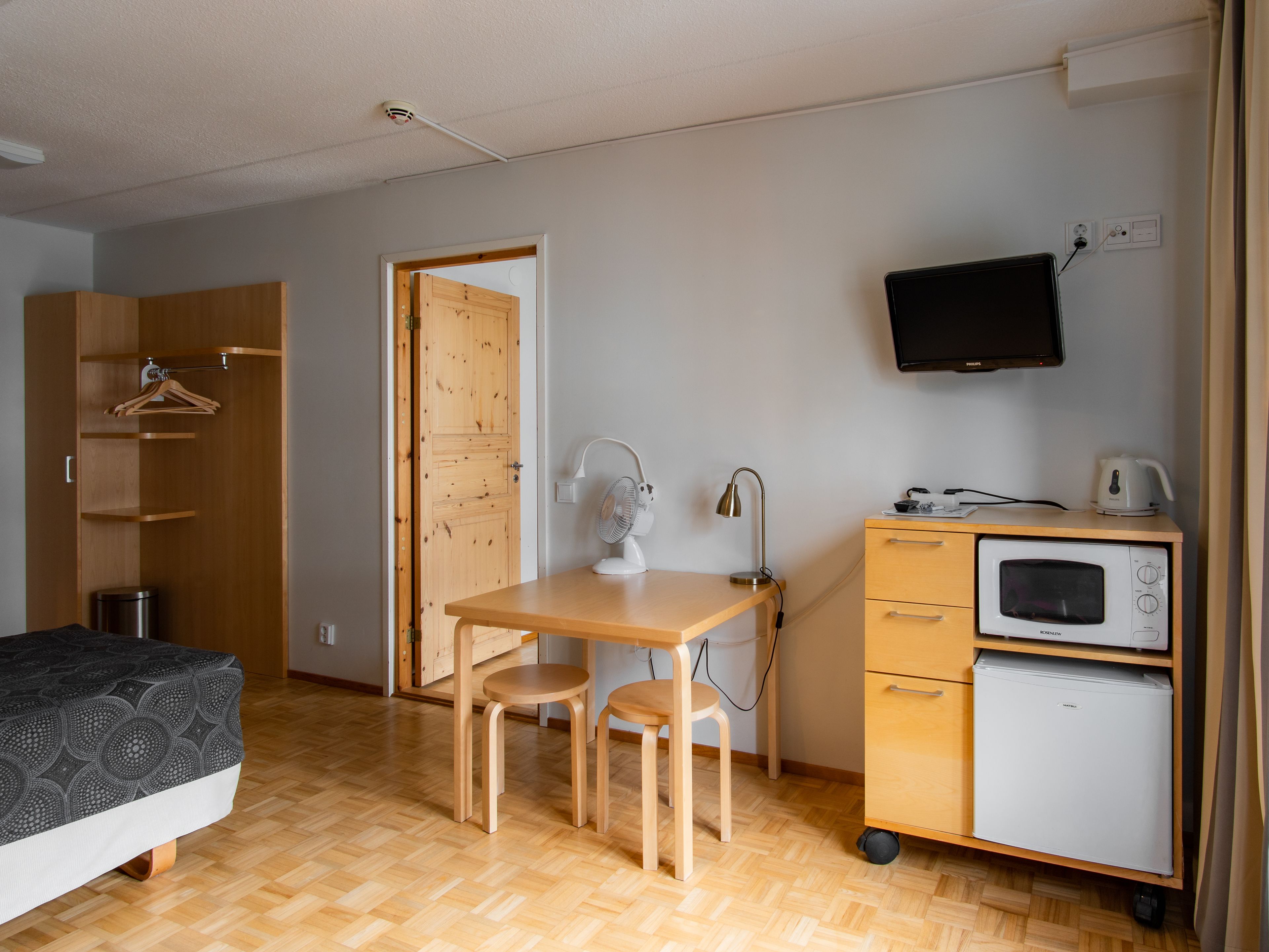 A compact room with a bed, a wooden desk with two chairs, a small kitchenette with a microwave and a mini-fridge, and a TV mounted on the wall.