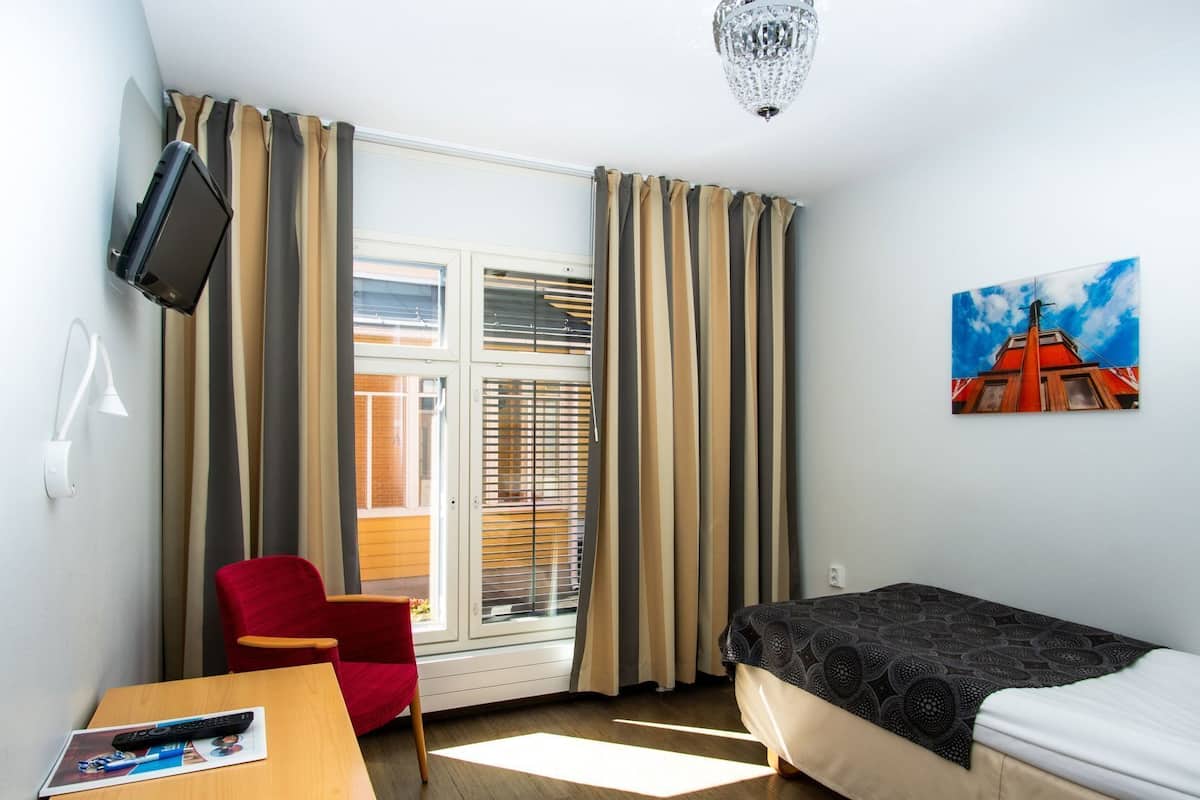 Ein Zimmer mit einem Bett, einem Schreibtisch mit Fernbedienung, einem roten Sessel, einem an der Wand hängenden Fernseher und einem Gemälde eines Gebäudes.