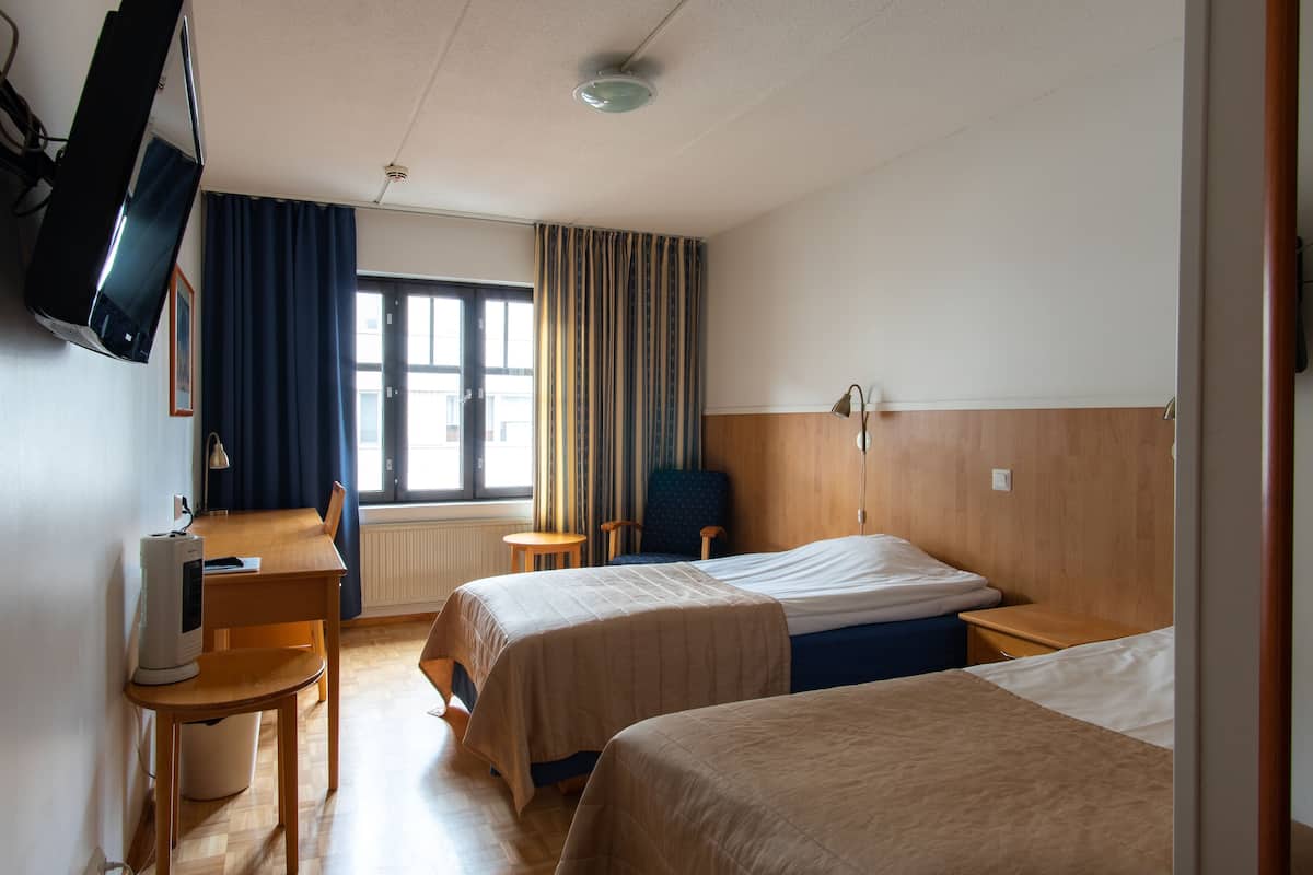 Ein Hotelzimmer mit zwei Betten, einem Schreibtisch mit Computer, einem an der Wand befestigten Fernseher und einem Fenster mit Vorhängen.