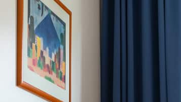 A room with a framed abstract painting on the wall, a desk with a lamp, and a window with dark curtains.