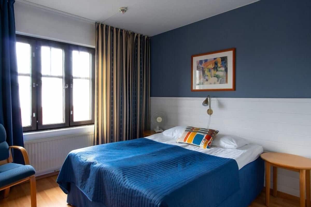 Ein Hotelzimmer mit Bett, Sessel, Nachttisch, Fenster mit Vorhängen und einem Bild an der Wand.