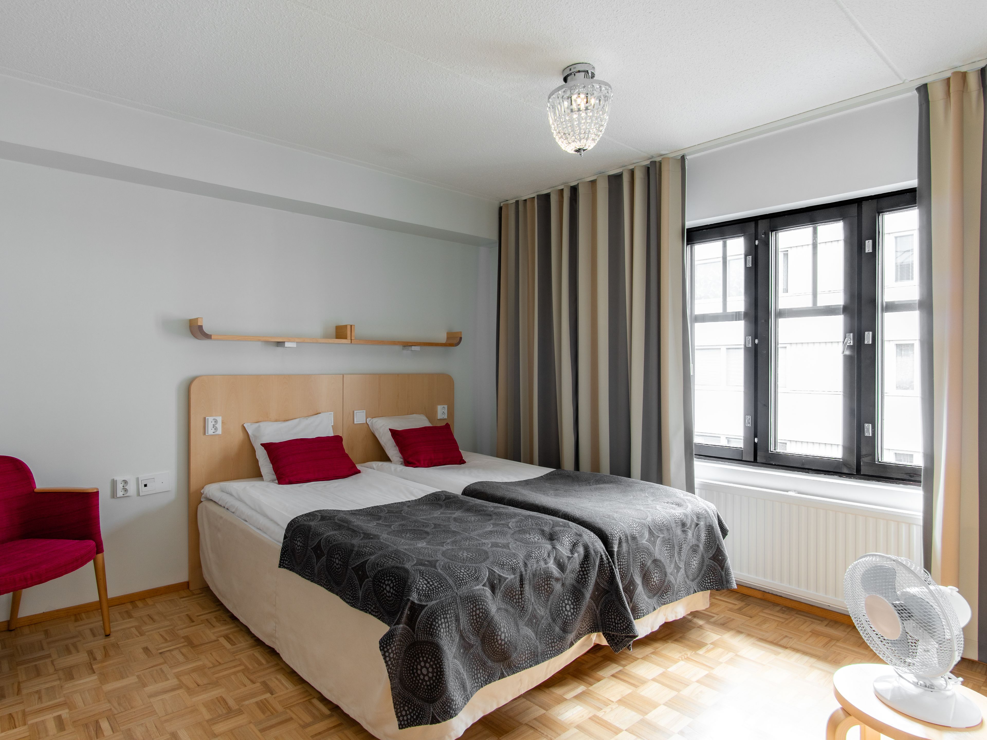 Ein Doppelbett mit Holzvorrahmen, zwei rote Kissen, eine graue, gemusterte Tagesdecke, ein Standventilator, ein roter Sessel und ein Fenster mit Rollläden.