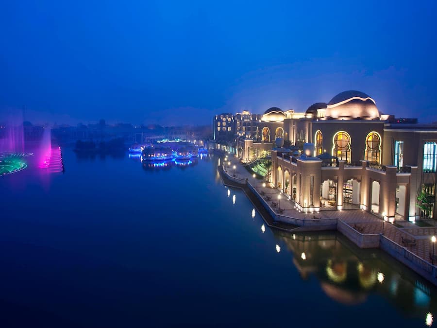 A waterfront hotel with a reflective pool, illuminated by colorful lights and a fountain.