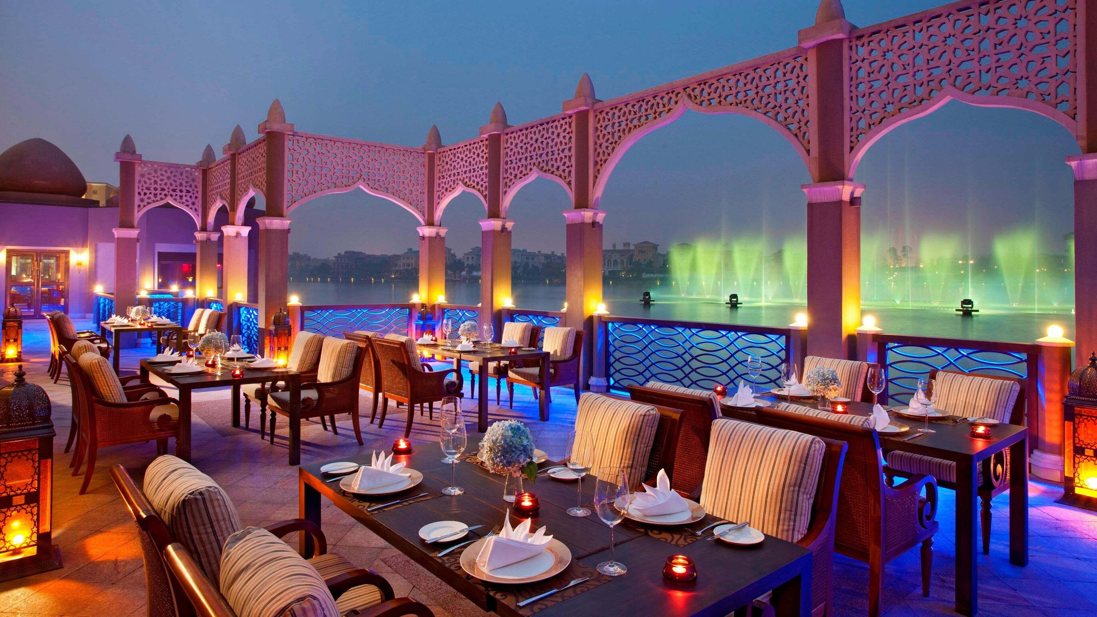 A rooftop dining area with tables set for dinner, overlooking a body of water with illuminated fountains.