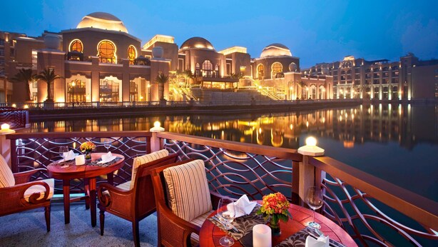 A waterfront dining area with a table set for two, overlooking a reflective body of water and a large building with domed structures.
