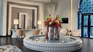 A modern lobby with a central round table, seating, a chandelier, and a large window.