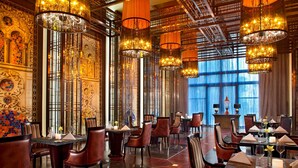 A restaurant with high ceilings, ornate chandeliers, and patterned walls. Tables are set with white napkins and candles.
