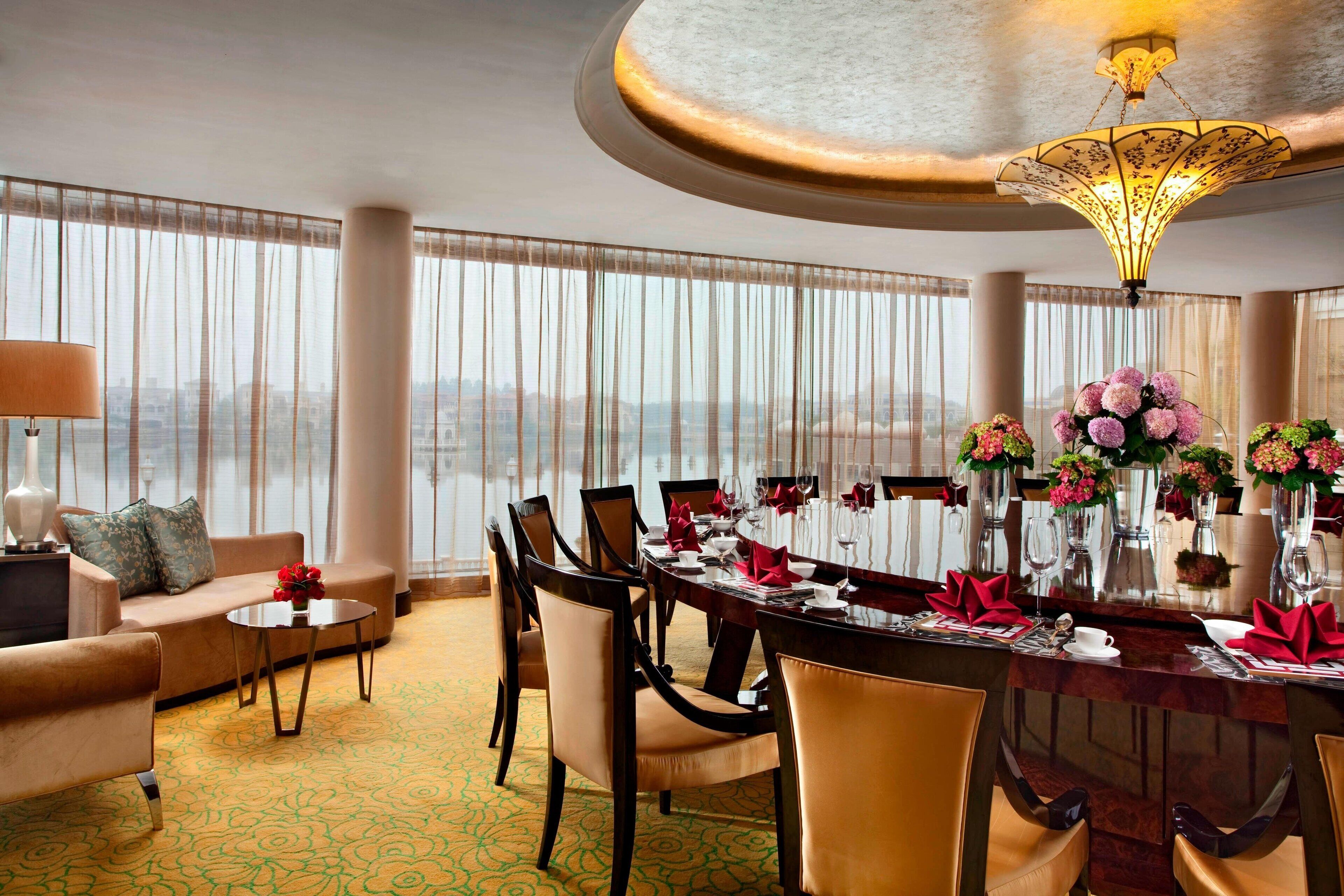 A hotel dining area with a round table set for a meal, surrounded by chairs, a sofa, and a lamp.