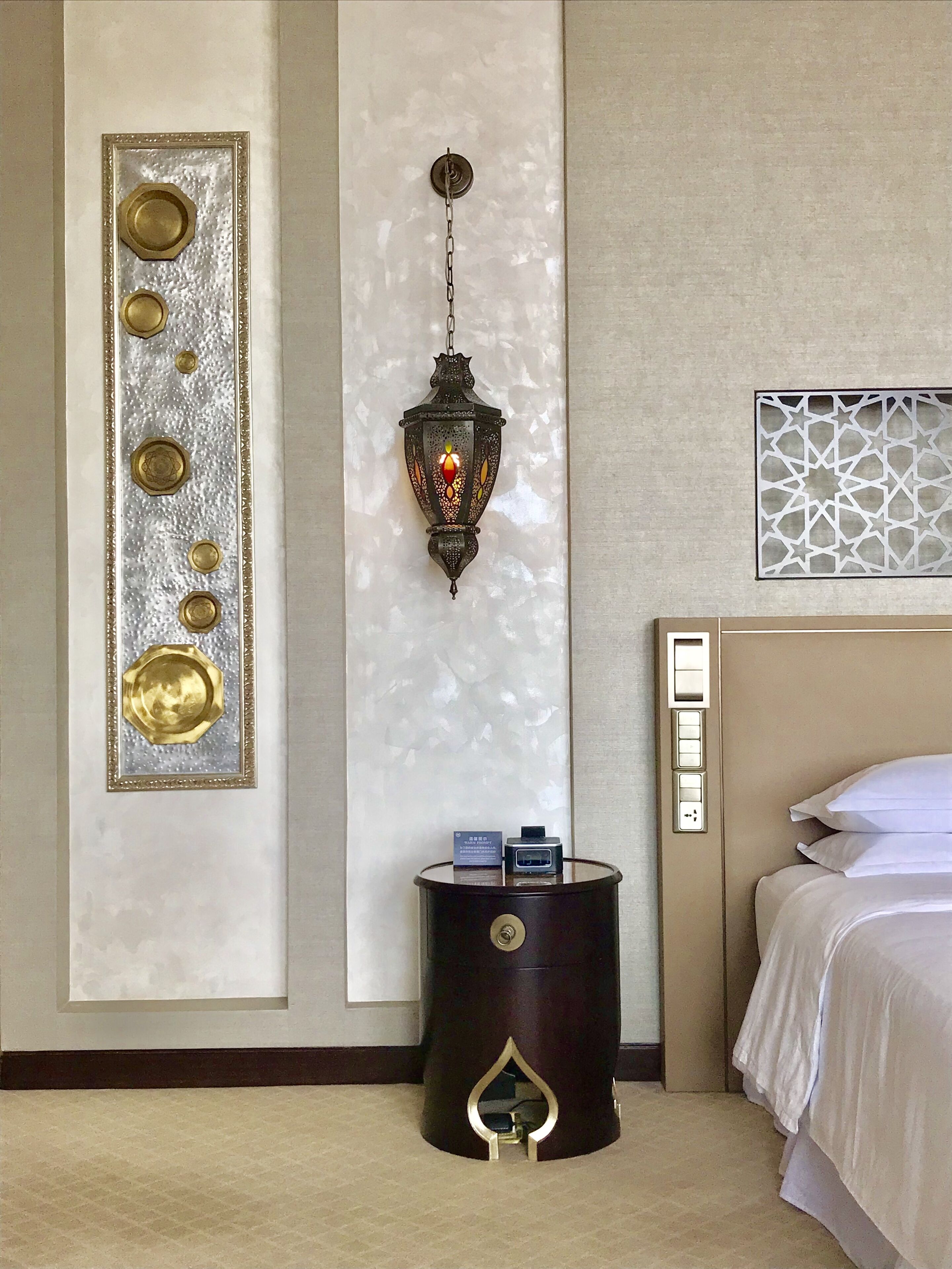 A hotel room with a bed, a wall-mounted decorative panel, a wall-mounted lantern, a small barrel with a lock, and a power outlet.