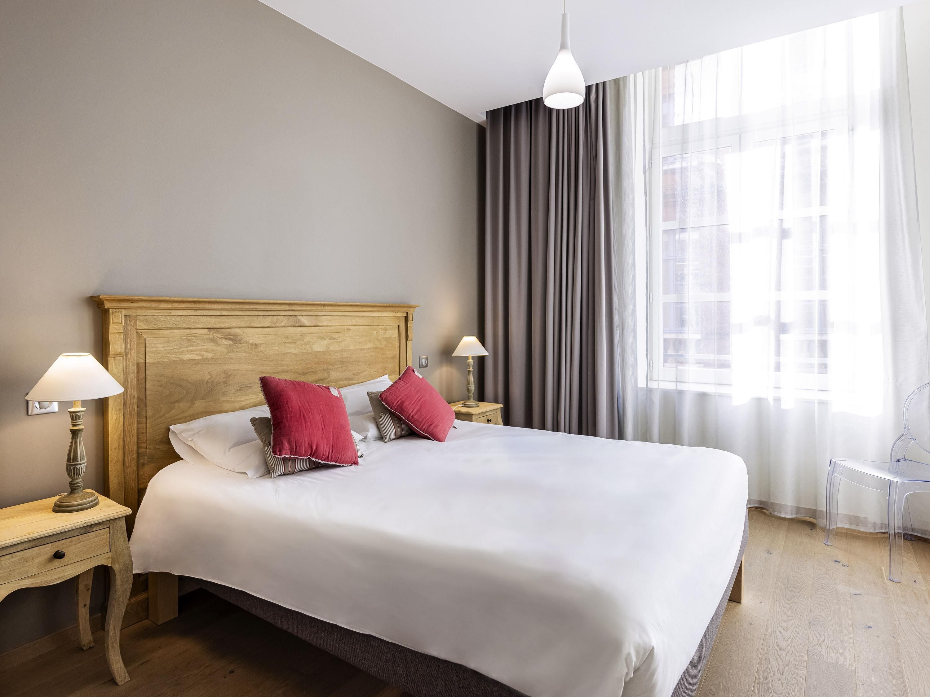 A neatly made bed with white linens and two red pillows, a wooden headboard, bedside lamps, and a clear chair by a window with curtains.