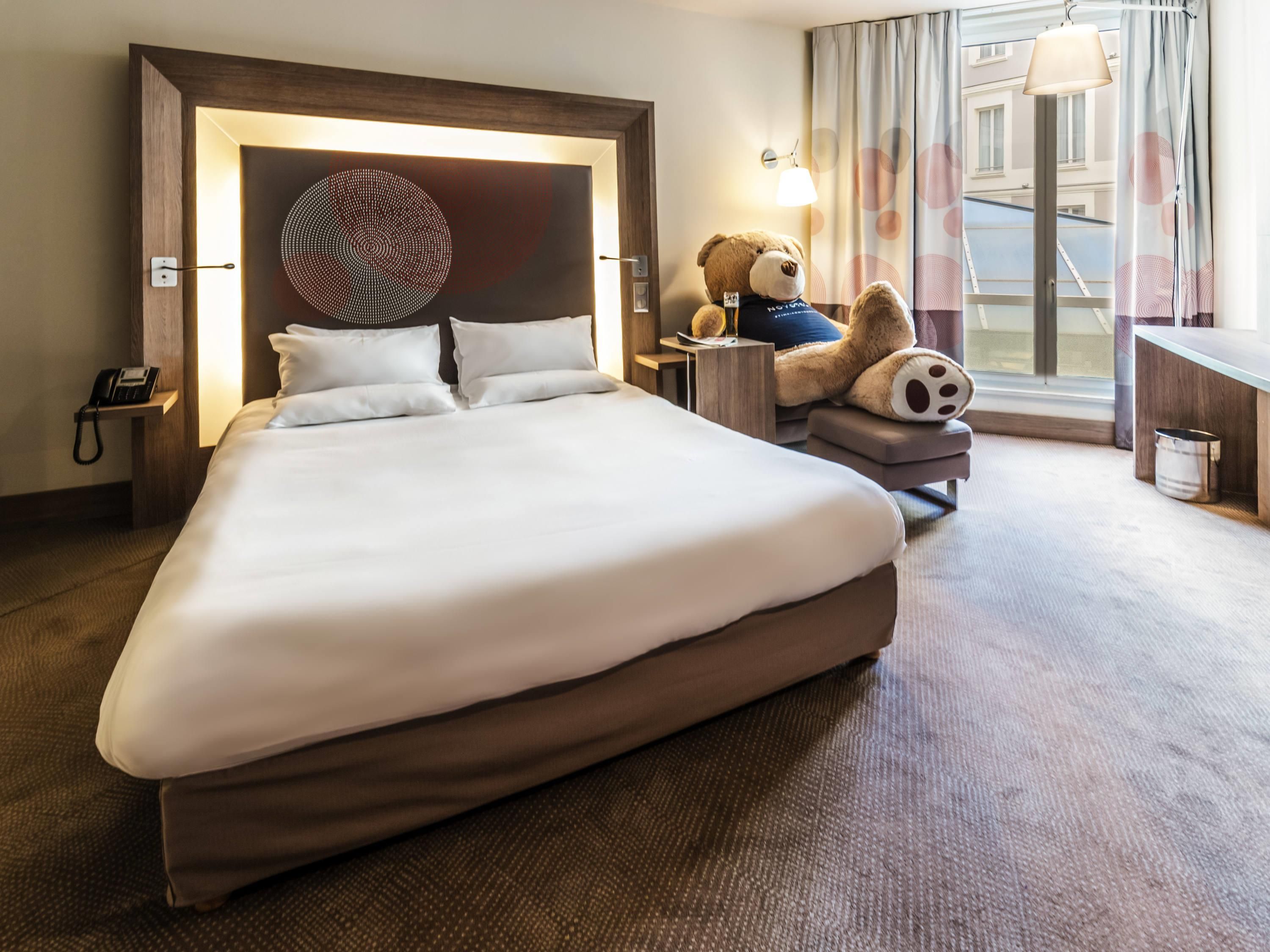 Habitación de hotel con una cama grande, una lámpara de noche, un oso de peluche y un escritorio con lámpara.