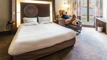 Habitación de hotel con una cama grande, una lámpara de noche, un oso de peluche y un escritorio con lámpara.