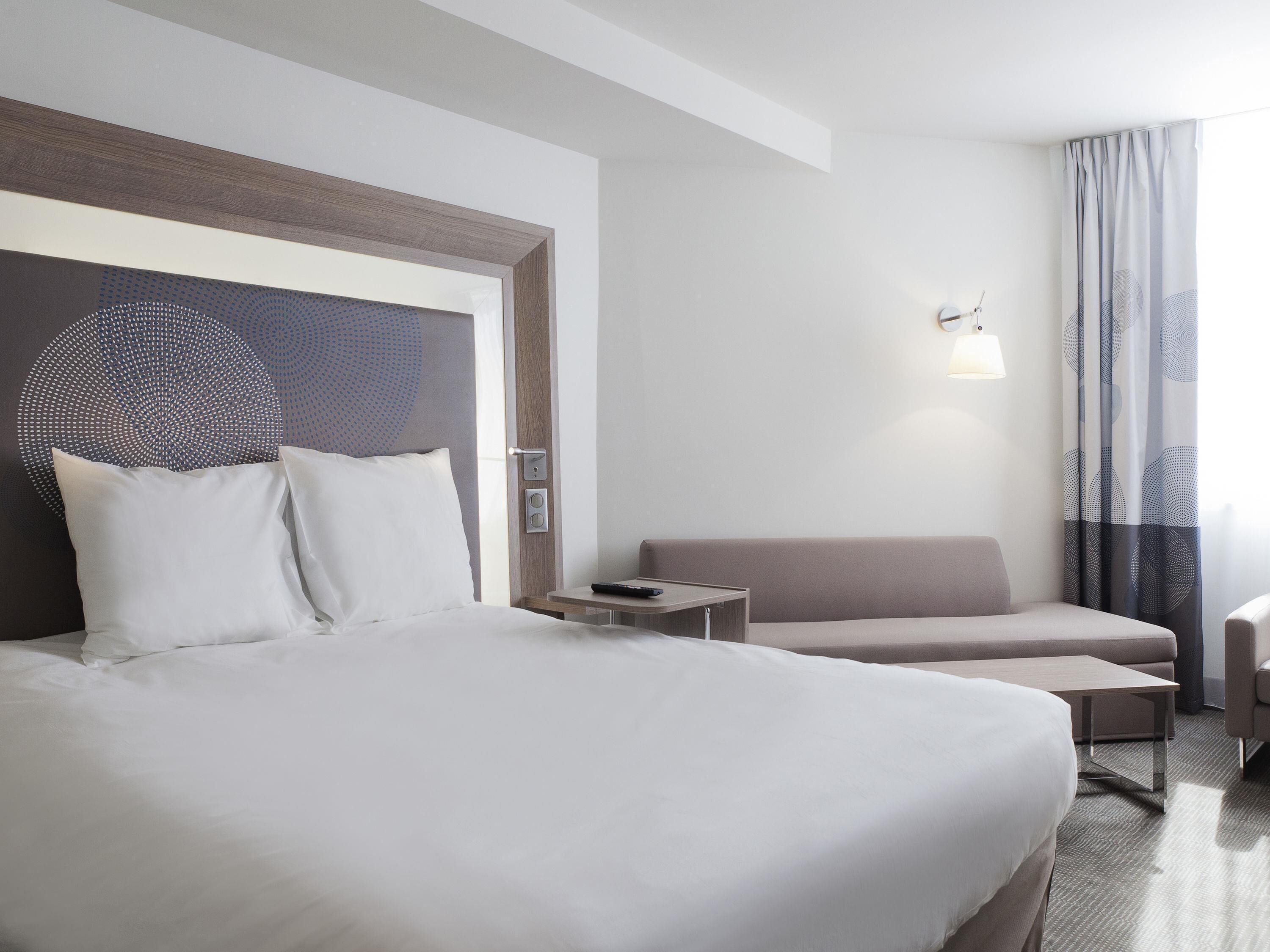 Habitación de hotel con una cama grande, un pequeño sofá, una mesita de noche y una lámpara de pared.