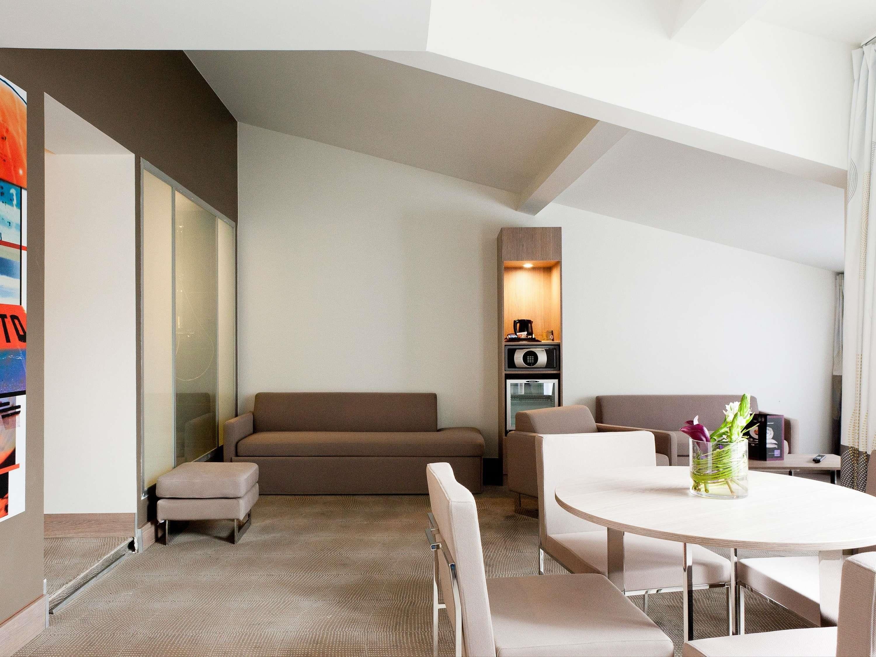 A modern hotel room with a sofa, a small table, and a kitchenette area.