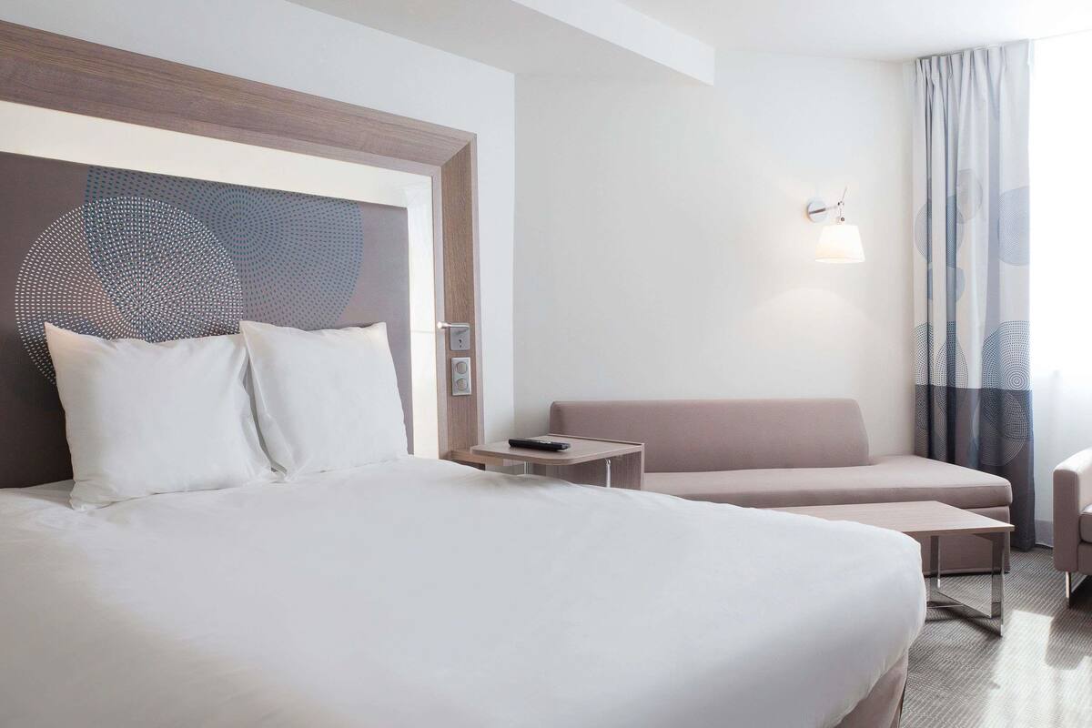 A hotel room with a large bed, a sofa, a small table, and a wall-mounted light fixture.