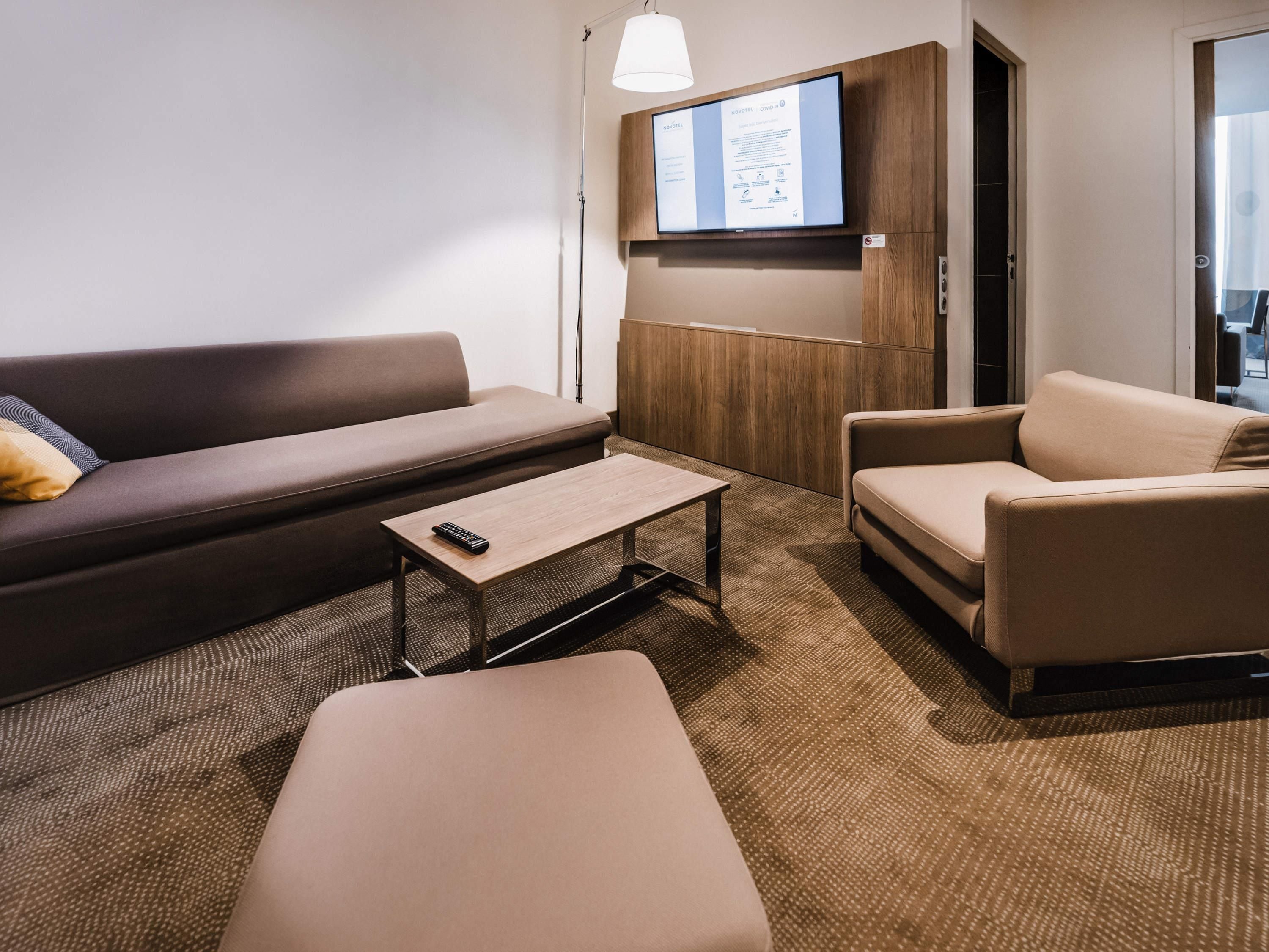Une chambre d’hôtel moderne équipée d’un canapé, d’un fauteuil et d’un pouf, d’un téléviseur à écran plat fixé sur un panneau en bois et d’une lampe.