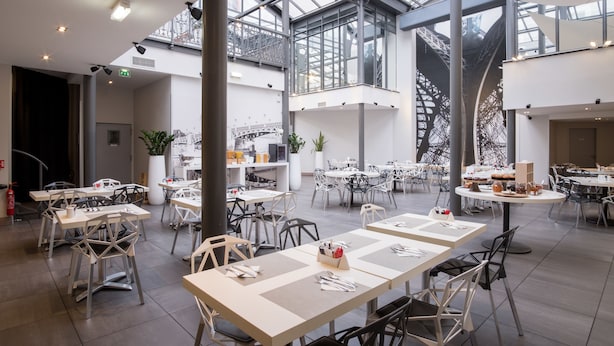 Un vaste espace de restauration intérieur, doté de hauts plafonds et de grandes fenêtres, aménagé avec un mobilier moderne et agrémenté d’un arrangement floral central.