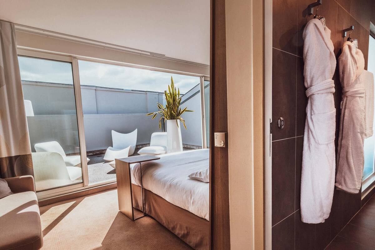 A modern hotel room with a large bed, a seating area with chairs, a potted plant, and a view of the sky.