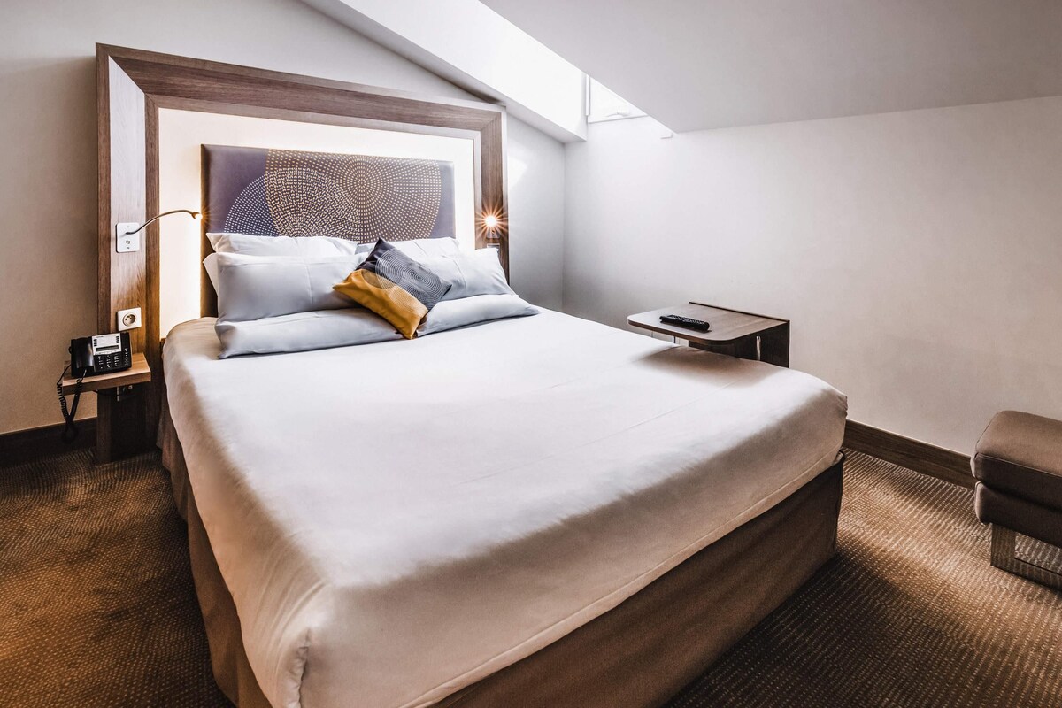 Ein Hotelzimmer mit einem großen Bett, einem Nachttisch mit Telefon, Fernbedienung und einem kleinen Sessel.