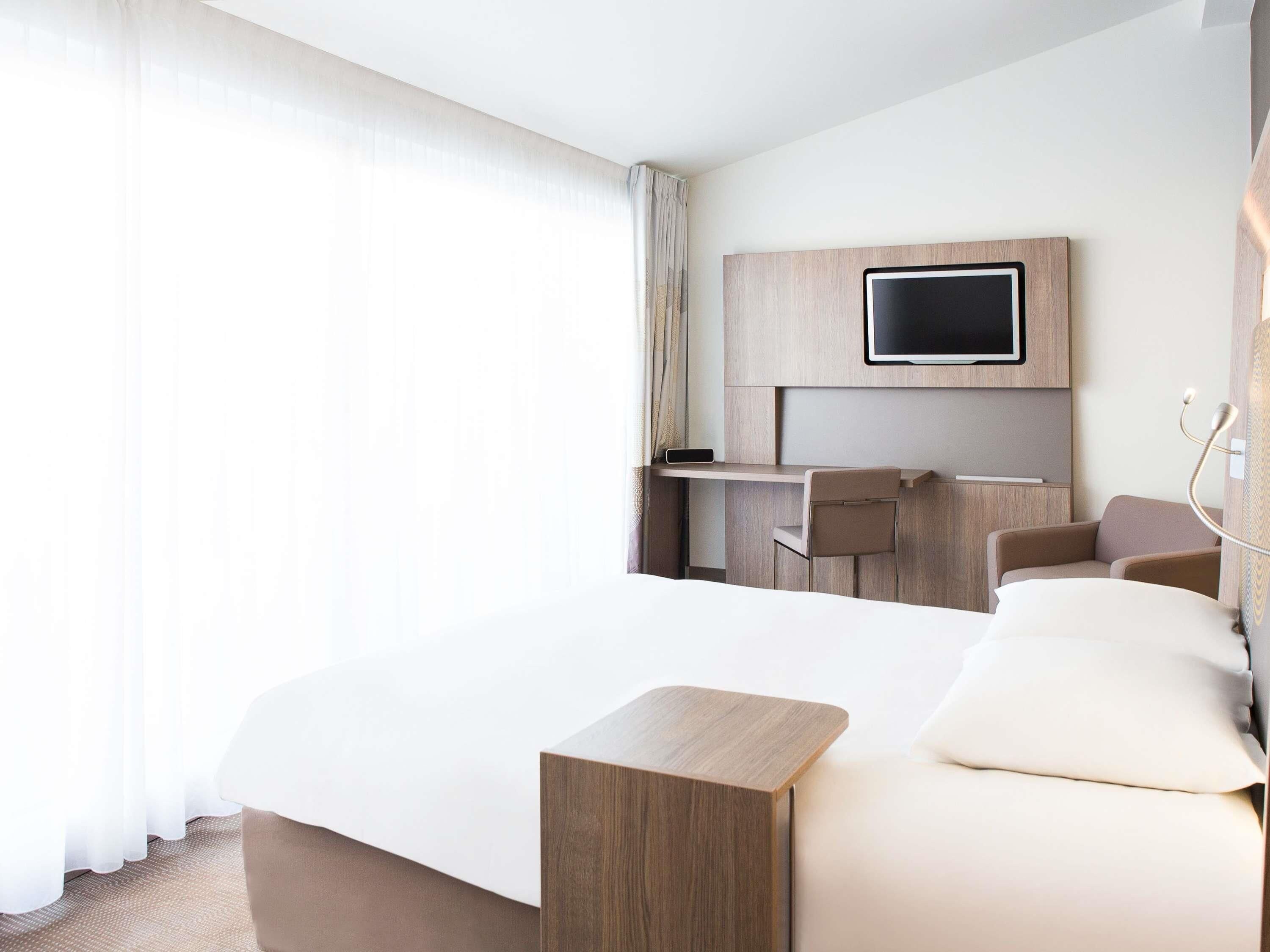 Quarto de hotel com cama, escrivaninha, cadeira, televisão e mesa de cabeceira.