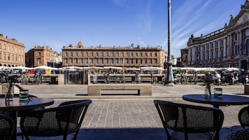 Un espace café en plein air avec des chaises et des tables en osier, une fontaine et, en arrière-plan, un bâtiment historique.