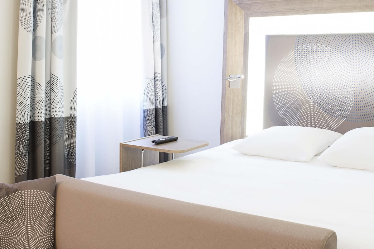 Een hotelkamer met een bed, een nachtkastje, een afstandsbediening en een raam met gordijnen.