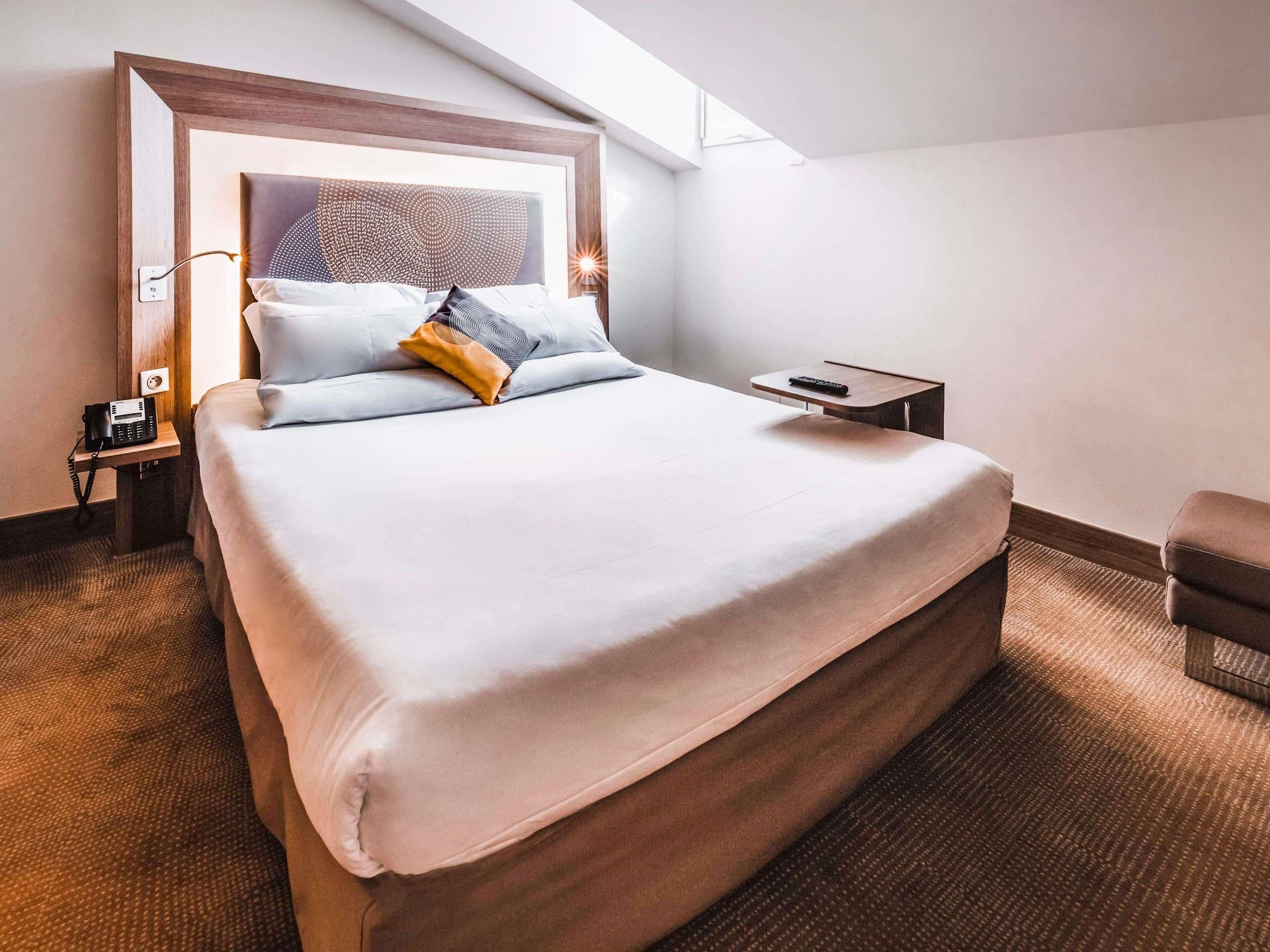 A hotel room with a large bed, a bedside table with a phone, a remote control, and a small chair.