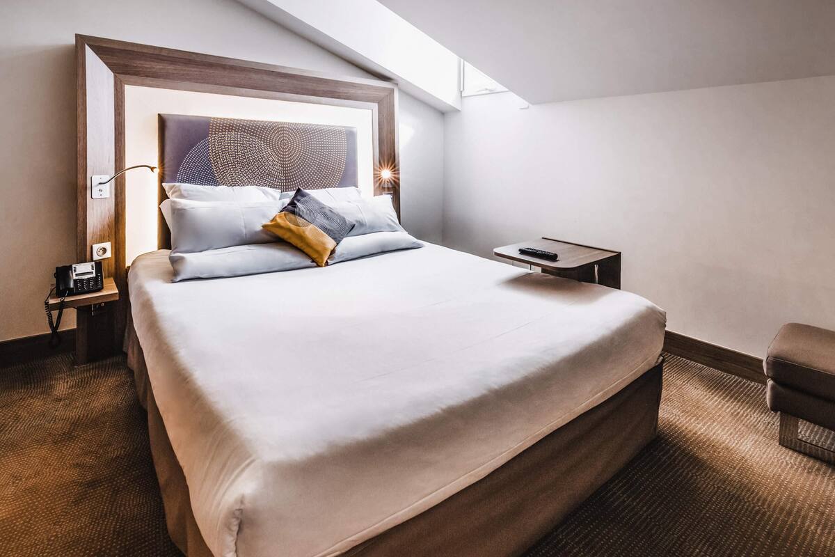 A hotel room with a large bed, a bedside table with a phone, a remote control, and a small chair.