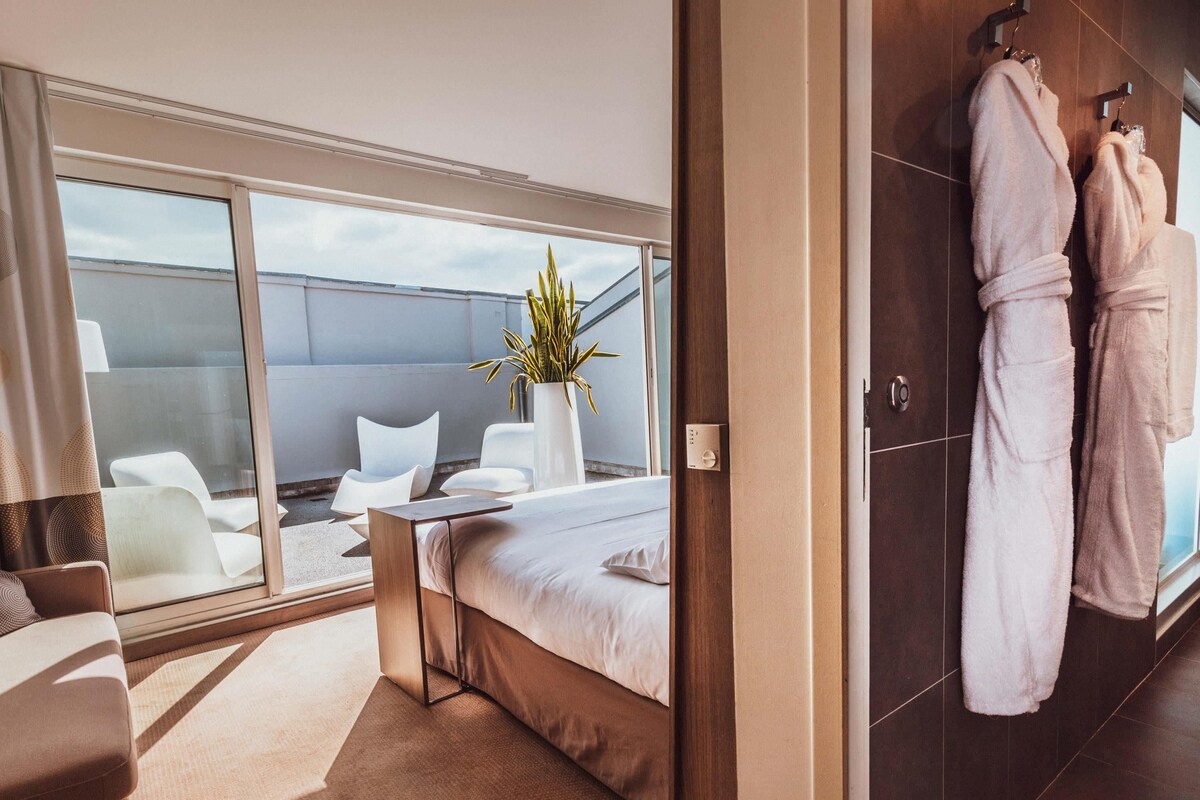 Ein modernes Hotelzimmer mit einem großen Bett, einer Sitzecke mit Stühlen, einer Topfpflanze und Blick zum Himmel.