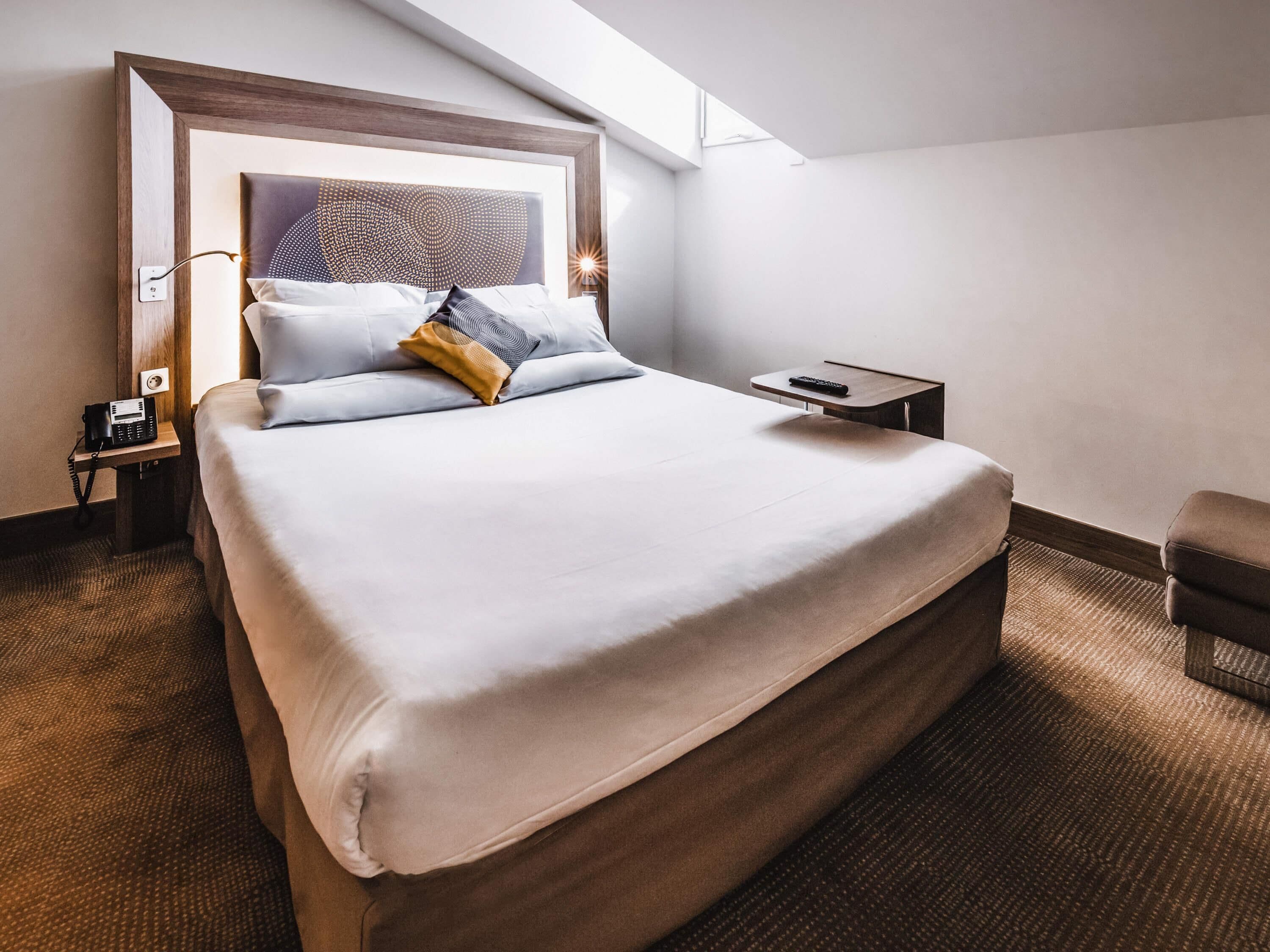 A hotel room with a large bed, a bedside table with a phone, a remote control, and a small chair.