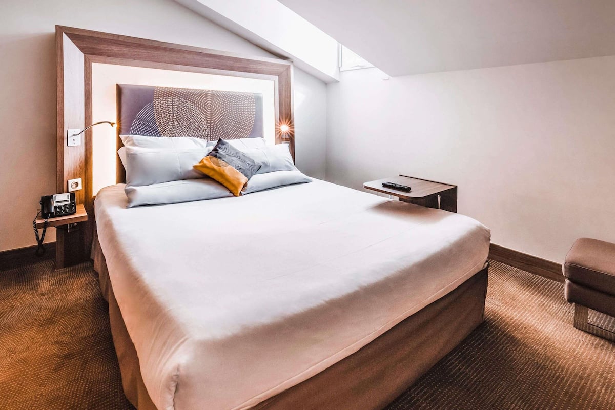 Hotelkamer met een groot bed, nachtkastje met telefoon, afstandsbediening en een kleine stoel.