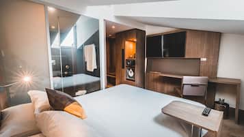 Habitación de hotel moderna con una cama grande, un escritorio, un televisor y un baño visible a través de un panel de vidrio.