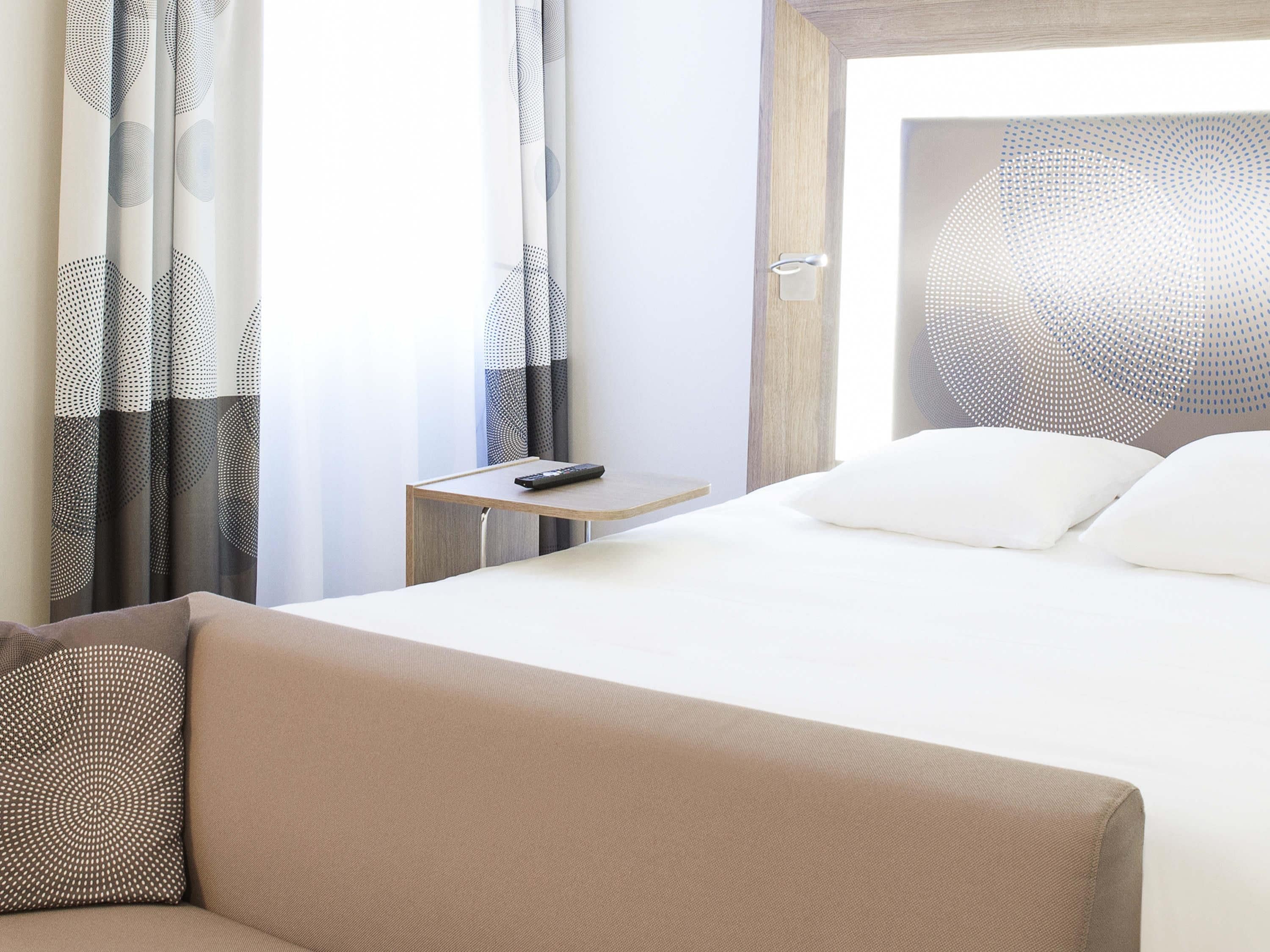 A hotel room with a bed, a nightstand, a remote control, and a window with curtains.