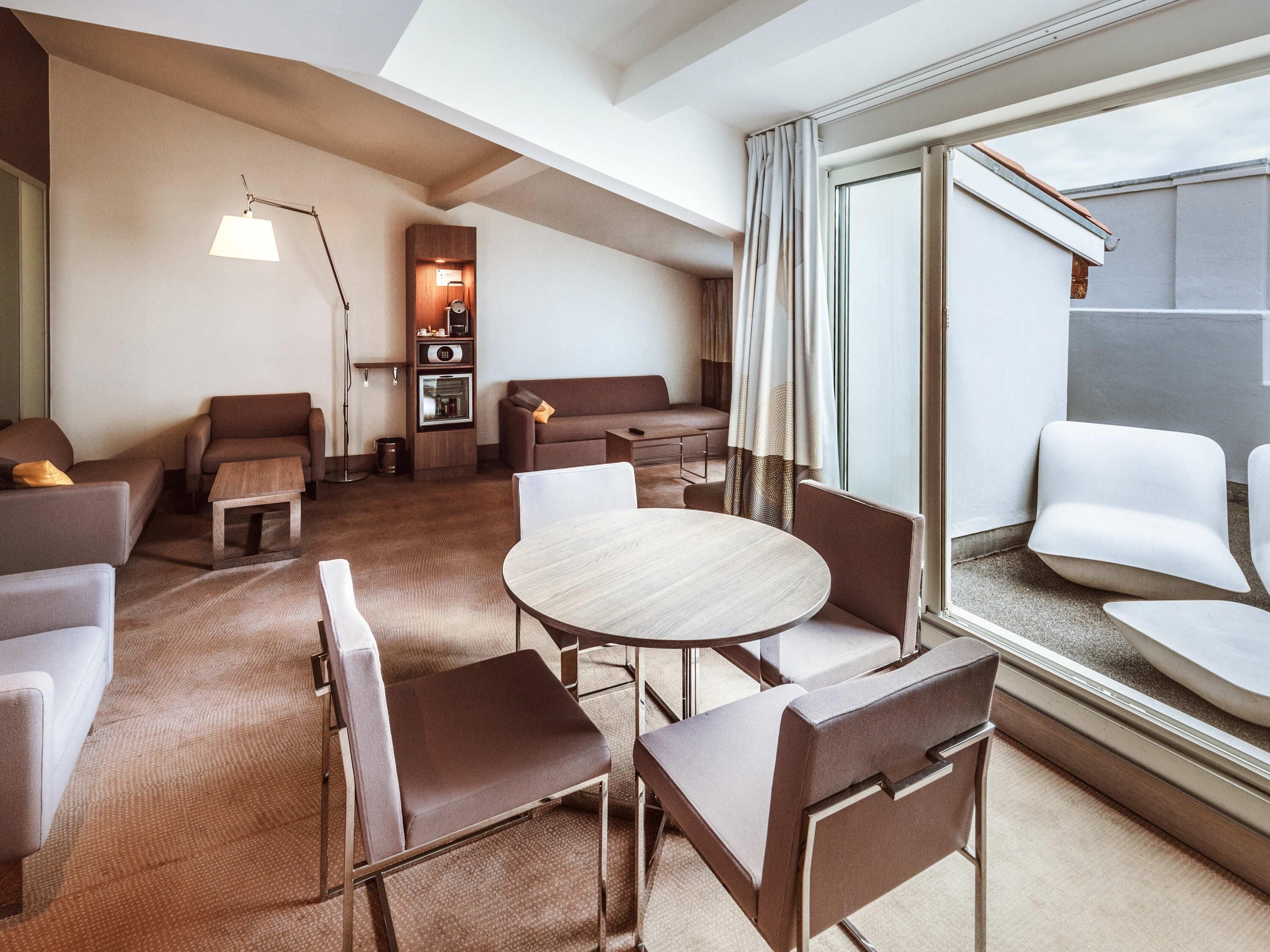 A modern hotel room with a round table and chairs, a sofa, a small cabinet, and a large window with curtains.