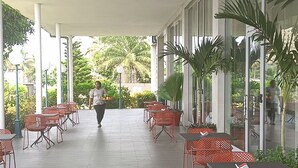 Un espacio al aire libre cubierto con filas de sillas y mesas rojas, ventanas amplias y plantas en macetas.