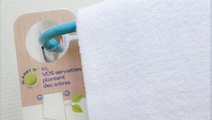 Etiqueta adherida a una toalla blanca con texto en varios idiomas que invita a reutilizar las toallas para plantar árboles.