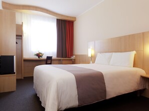 A hotel room with a large bed, a desk with a chair, a TV, and a window with curtains. - ibis Bata (Bata)
