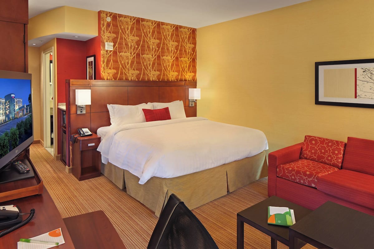 A hotel room with a large bed, a red sofa, a desk with a computer, a TV, and a wall-mounted artwork.