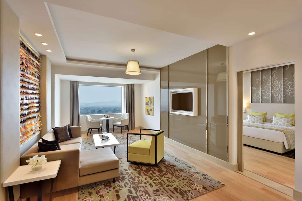 Ein modernes Hotelzimmer mit einer Couch, einem kleinen Tisch, einem gelben Hocker, einem großen Fenster mit Blick und einem Fernseher sowie einem Bett mit gemusterten Betttextilien.