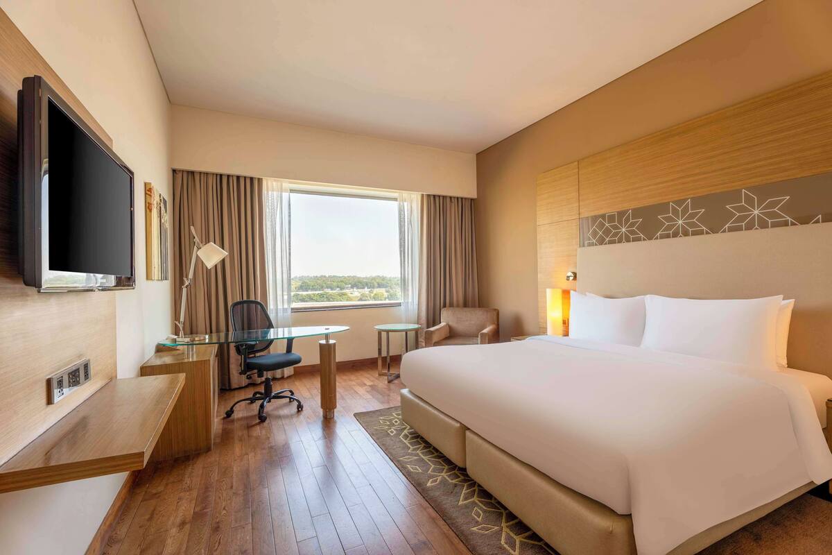 Ein Hotelzimmer mit einem großen Bett, einem Schreibtisch mit Stuhl, einem Fernseher und einem Fenster mit Vorhängen.