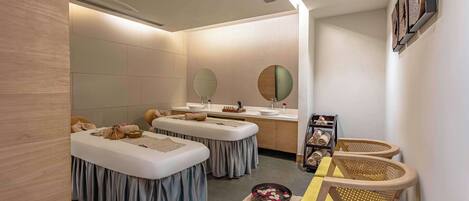 Un salón de spa con dos camillas de masaje, un lavamanos y un espejo. Hay sillas y un florero en el suelo.