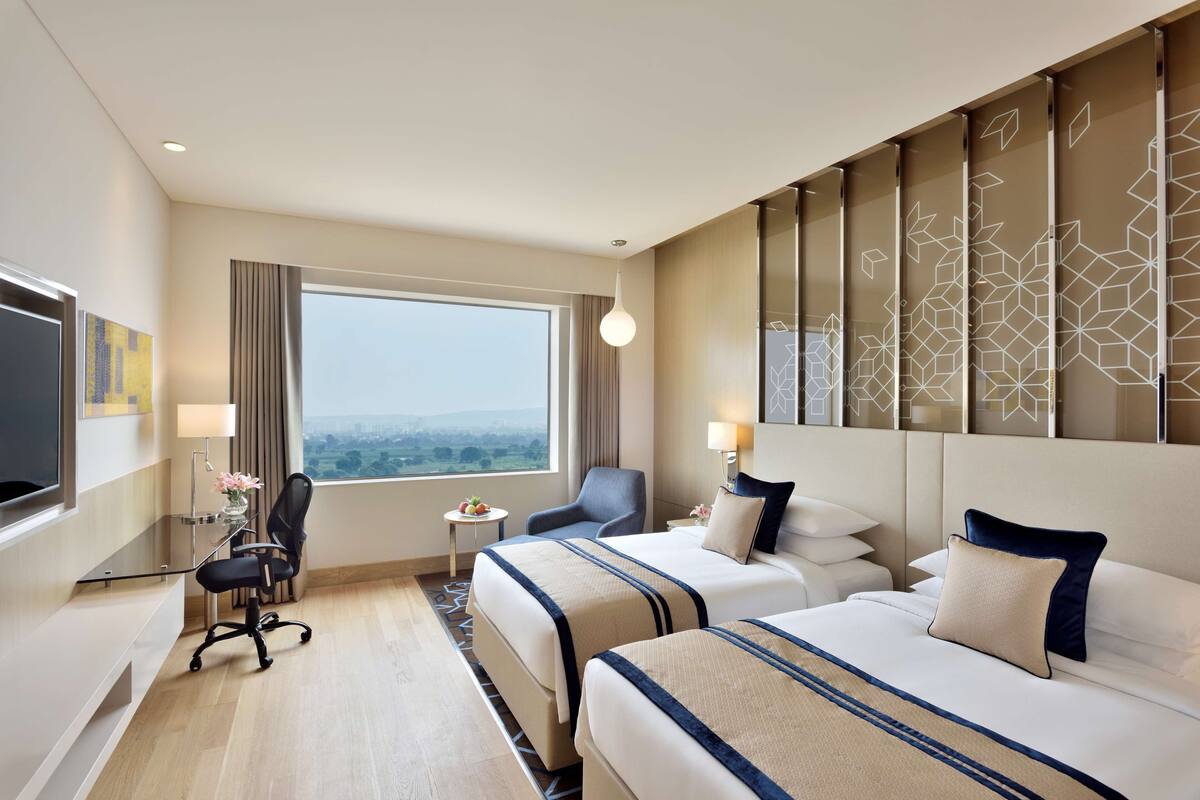 Ein Hotelzimmer mit zwei Betten, einem Schreibtisch, einem Stuhl, einem Fernseher und einem großen Fenster.