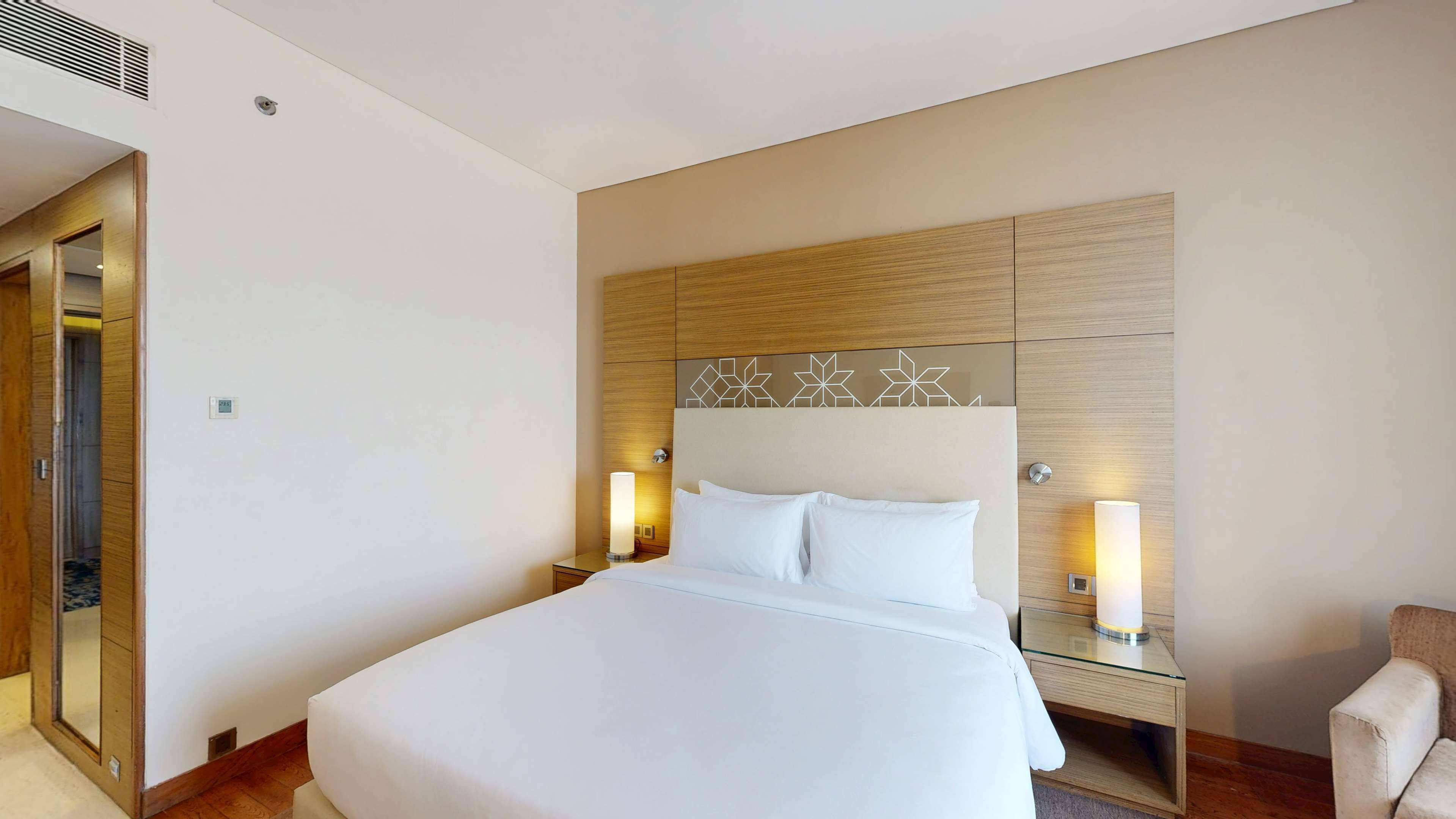 A modern hotel room with a large bed, two bedside tables with lamps, a wooden headboard with geometric patterns, and a mirror on the left wall.