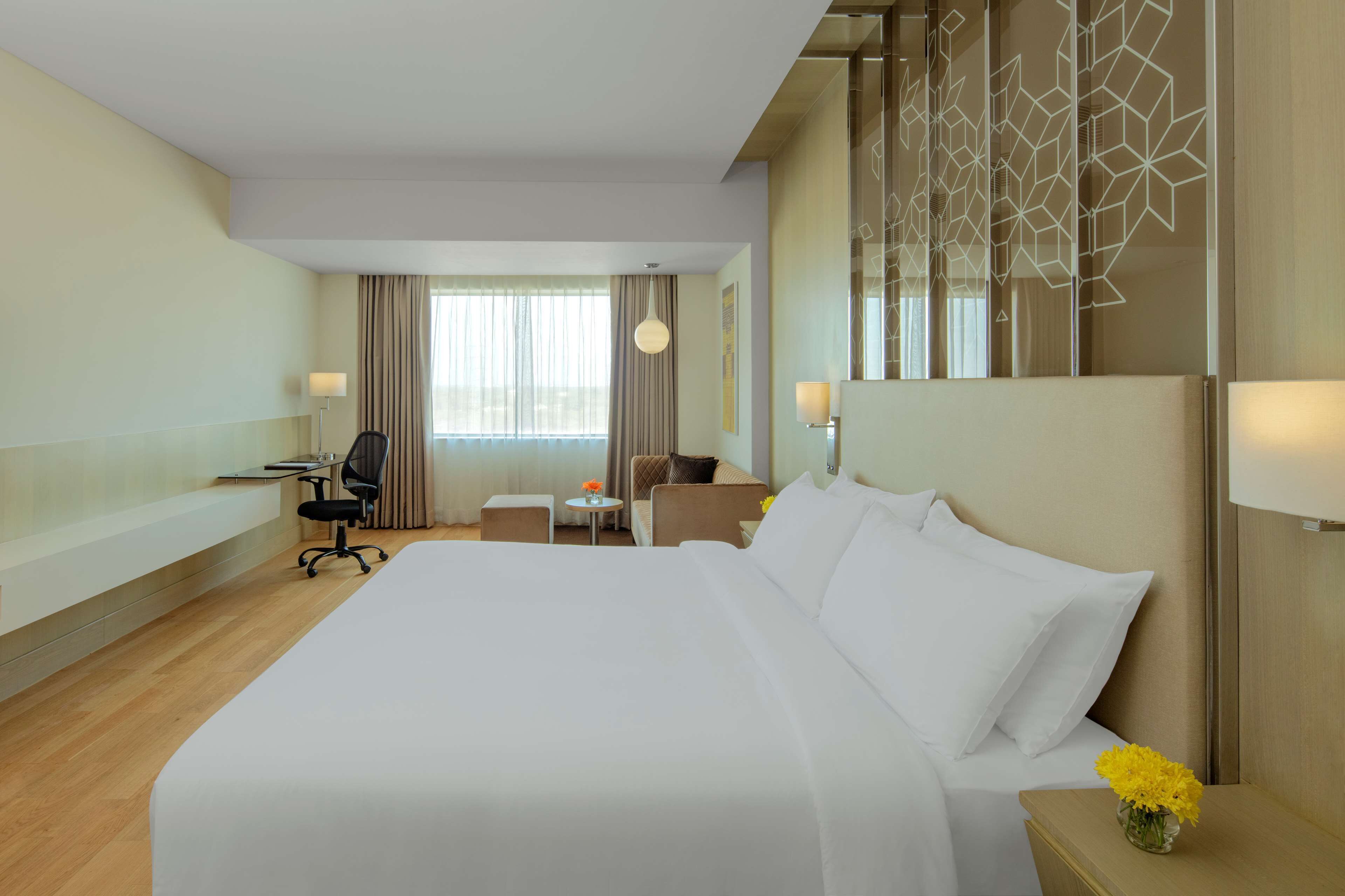 Een hotelkamer met een groot bed, een bureau, een stoel, een klein tafeltje met een bloevas, en uitzicht op het omliggende landschap door een raam.