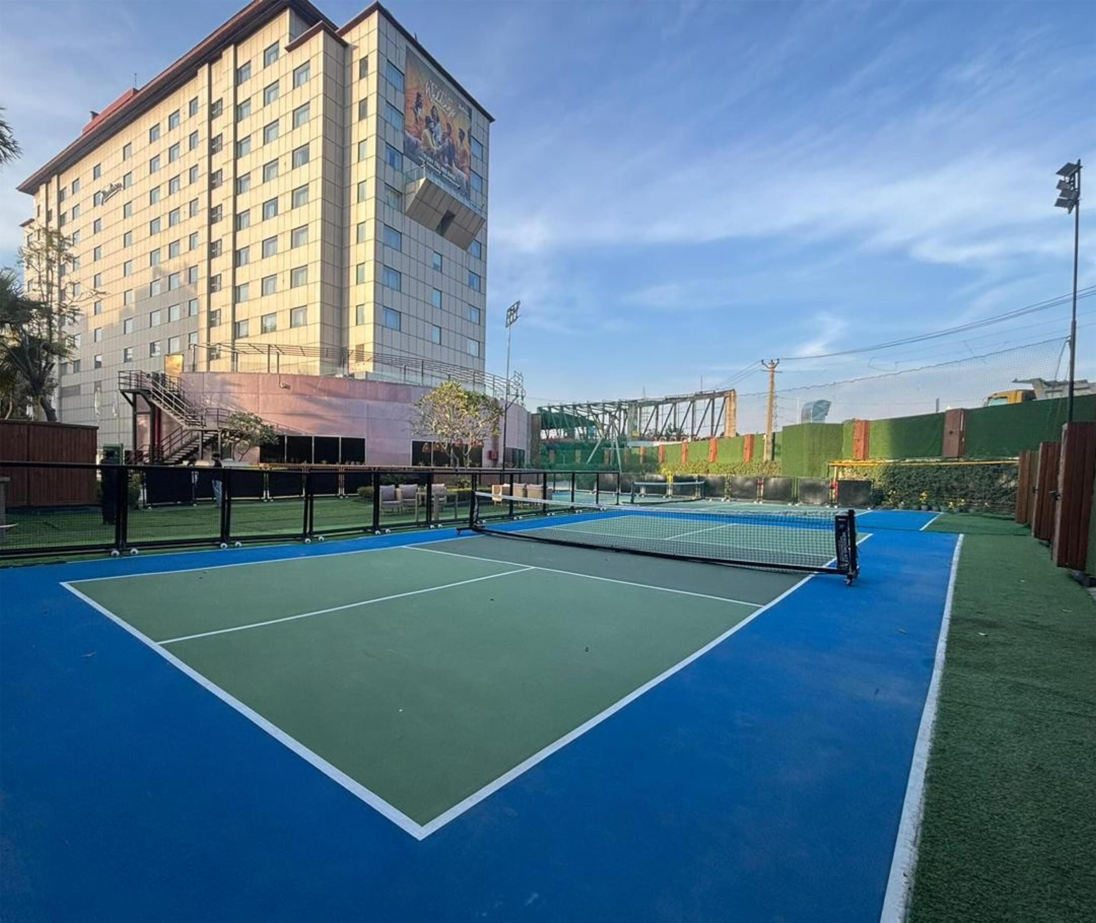 Een buitenterrein voor tennis met een net, omringd door een hek, en een hotelgebouw op de achtergrond.