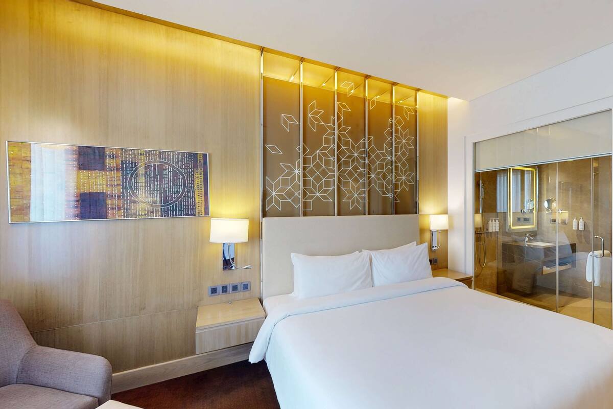 Ein modernes Hotelzimmer mit einem großen Bett, einer Sitzecke mit einem Sessel, einem Glaskastenbad und einer dekorativen Wandverkleidung.