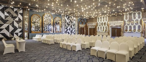 Un salón de conferencias con filas de sillas blancas, una alfombra estampada y un techo con numerosas luces.