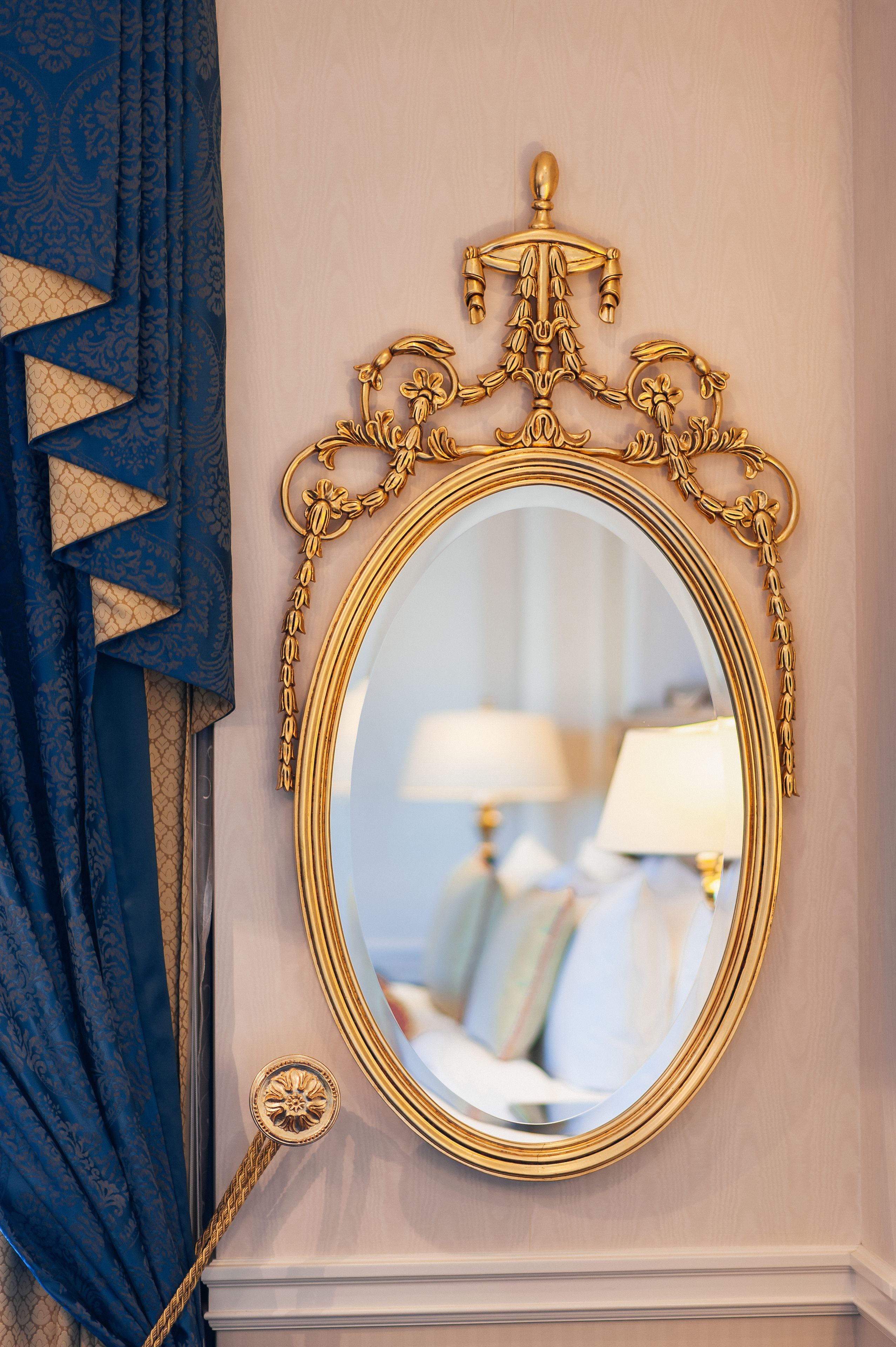 Espejo ornamentado con marco dorado y un atril decorativo, montado en una pared con una cortina azul estampada a la izquierda.